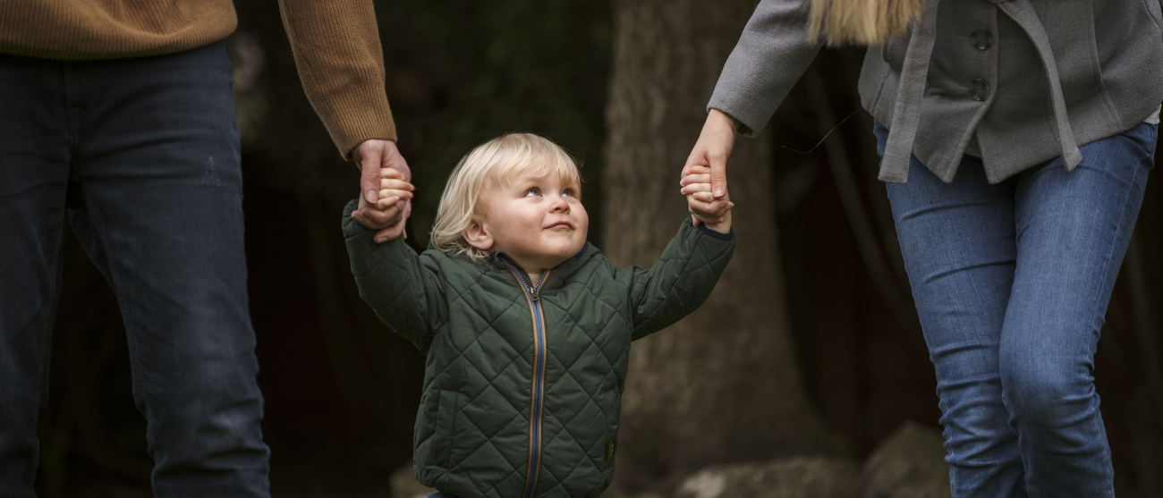 Dziecko w wieku przedszkolnym idzie z rodzicami za rączkę przez park.