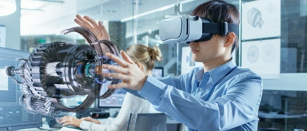 Mężczyzna w okularach VR pracuje z trójwymiarowym modelem holograficznym, wykonując gesty. W tle biuro inżynierskie oraz inni pracownicy.