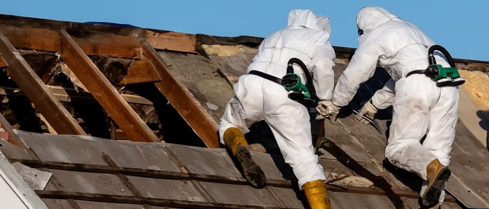 Dwóch ludzi w kombinezonach ściąga azbest z dachu.