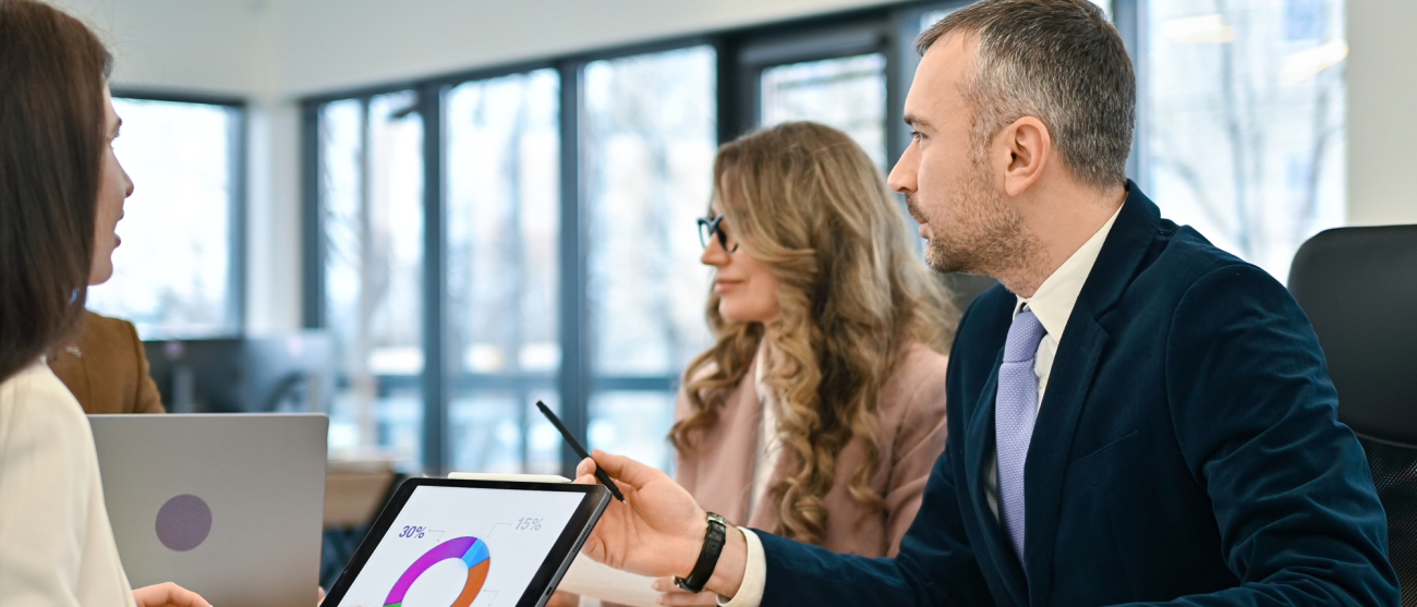 Zmiana Regulaminu projektów Kobieta i mężczyzna siedzą na spotkaniu a przed nim kolejna kobieta pracuje przy laptopie