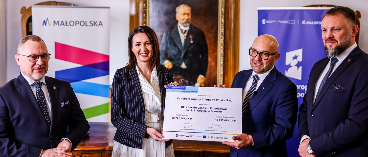 Grupa czterech elegancko ubranych osób stoi w reprezentacyjnym wnętrzu, prezentując duży certyfikat wsparcia finansowego dla Carlsberg Supply Company Polska S.A. W tle widoczne są banery promocyjne Małopolski i Funduszy Europejskich oraz portret historycznej postaci na ścianie.