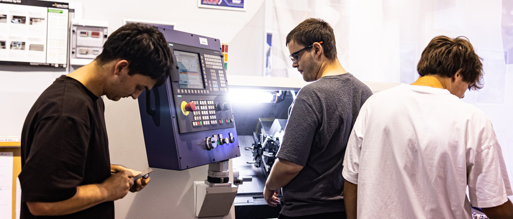 Trzech uczniów podczas zajęć praktycznych przy obrabiarce CNC w szkolnej pracowni technicznej